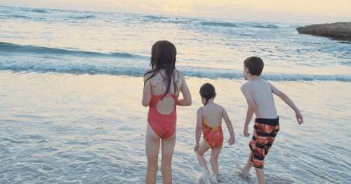 Três crianças na praia olhando para o horizonte em um cenário ensolarado.