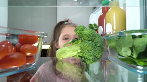 Uma menina abrindo a geladeira e pegando vegetais frescos.